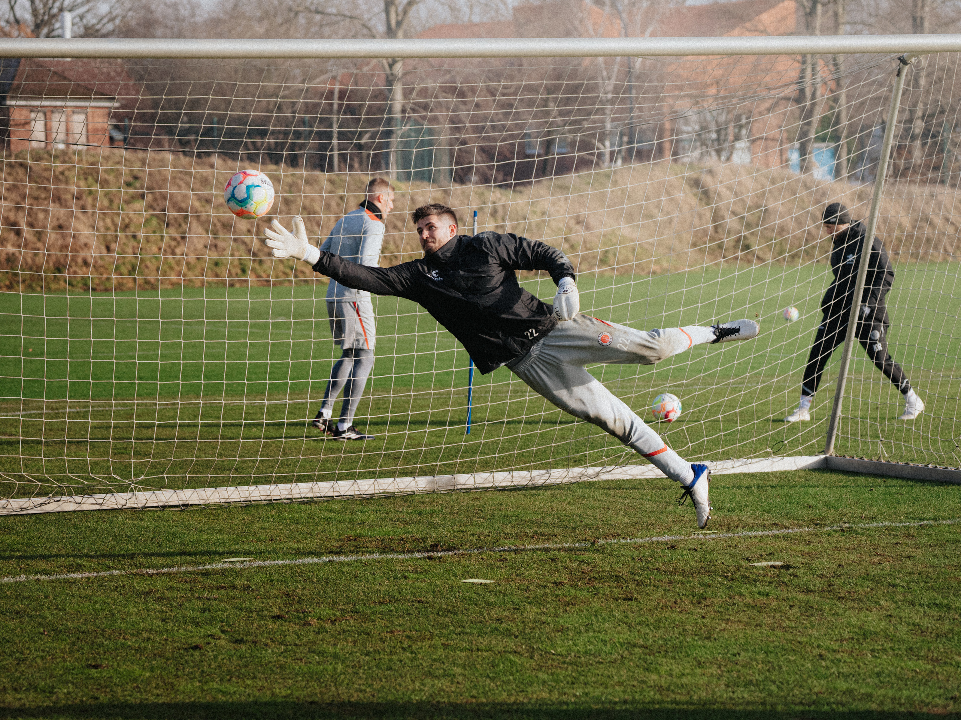 Nikola Vasilj springt beim Training einem Ball hinterher und streckt dabei den rechten Arm aus. Im Hintergrund sieht man ein Tor und zwei weitere St. Pauli-Spieler, die sich einen weiteren Ball zuspielen.