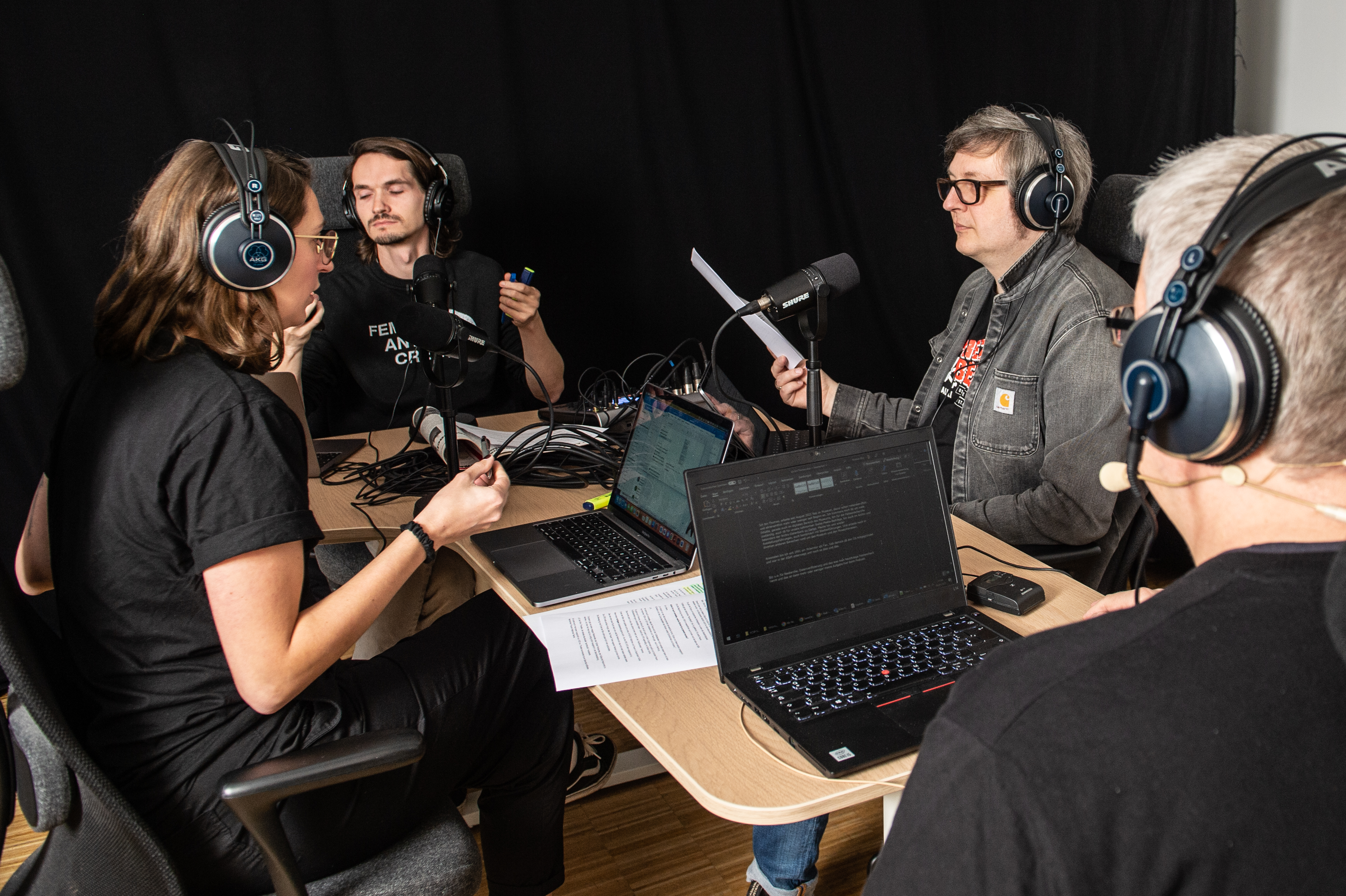Das Podcast-Team des Museums bei der Aufzeichnung der neuen Folge. Vier Personen sitzen sich an einem Tisch gegenüber, alle haben Kopfhörer auf und einen Computer vor sich auf dem Tisch stehen.